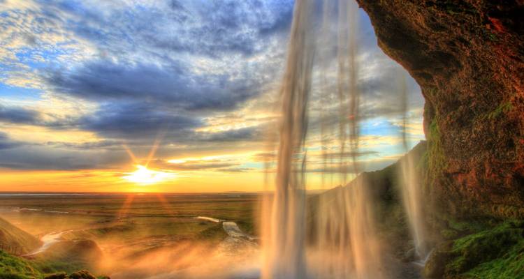 Die Sonne durchbricht den herabstürzenden Seljalandsfoss-Wasserfall und beleuchtet den Nebel über isländischen Ebenen bei Sonnenuntergang.