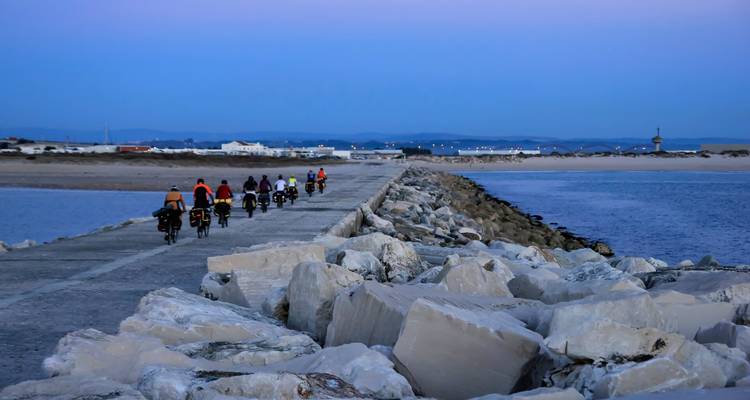 Line of cyclists ride along a long stone jetty flanked by boulders at dusk with coastal town lights ahead.