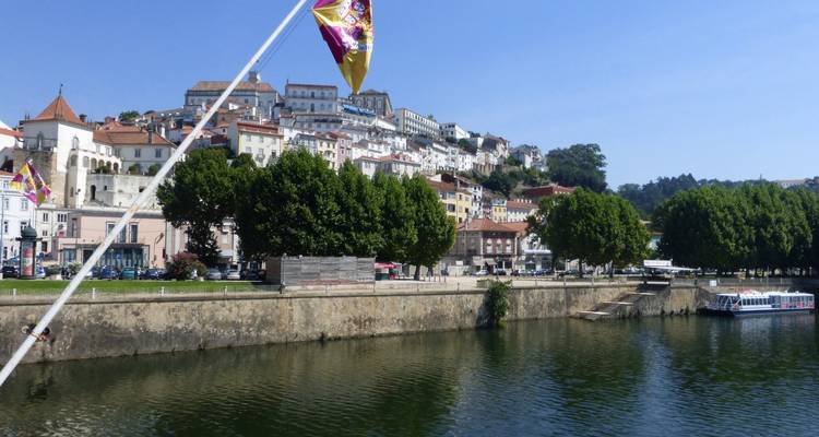 Riverfront view of Coimbra’s terraced white houses and university buildings rising on a hill under blue sky.