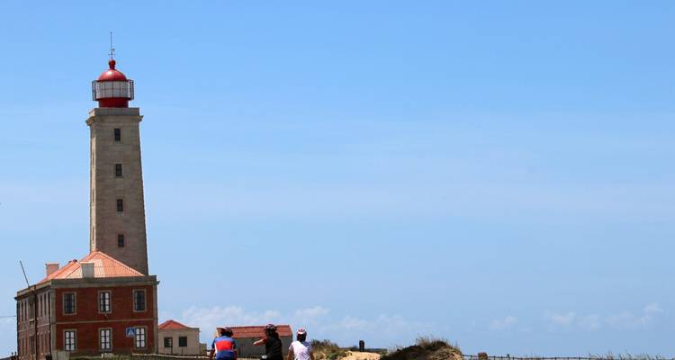 Tall coastal lighthouse with red lantern rises above low brick keeper’s house under cloudless sky while two tourists look on.
