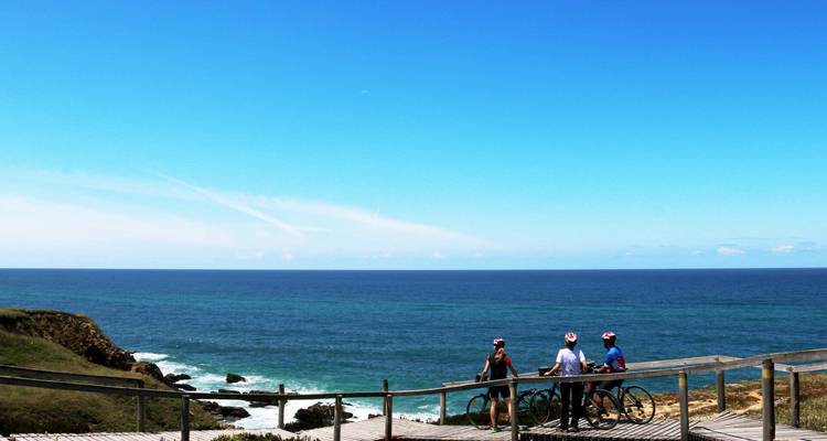 Three cyclists pause on a wooden deck overlooking the deep blue Atlantic under a cloudless sky.