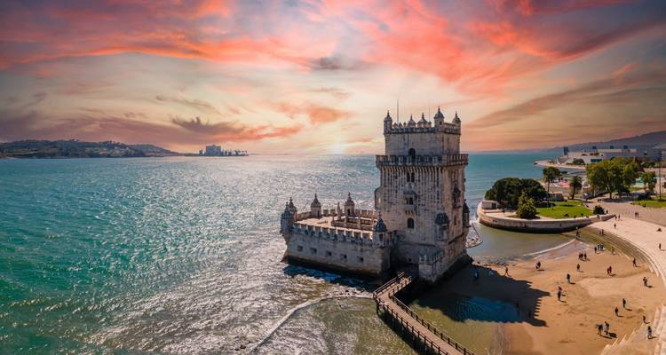Sunset sky paints vivid reds over Belém Tower jutting into the Tagus River with city shoreline in the background.
