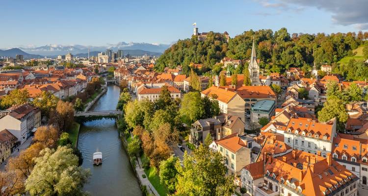 Une vue aérienne ensoleillée de Ljubljana montre la rivière, les ponts et la vieille ville aux toits rouges sous un ciel bleu.