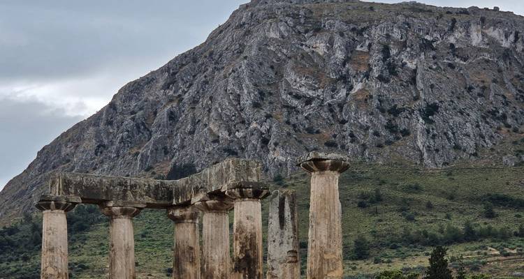 Columnas dóricas desgastadas de ruinas de templo antiguo situadas frente a una montaña escarpada de piedra caliza bajo un cielo gris.