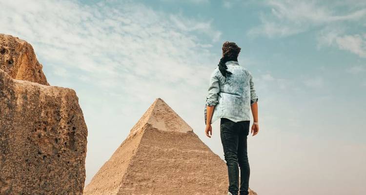 Un voyageur se tient sur des pierres anciennes contemplant une pyramide qui se dresse contre un ciel légèrement nuageux.