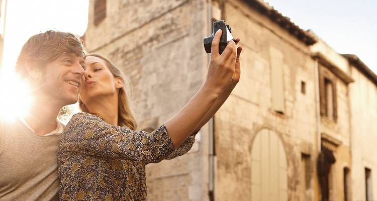 Pareja sonriente tomándose una selfie besándose bajo la cálida luz del sol junto a un antiguo edificio de piedra.