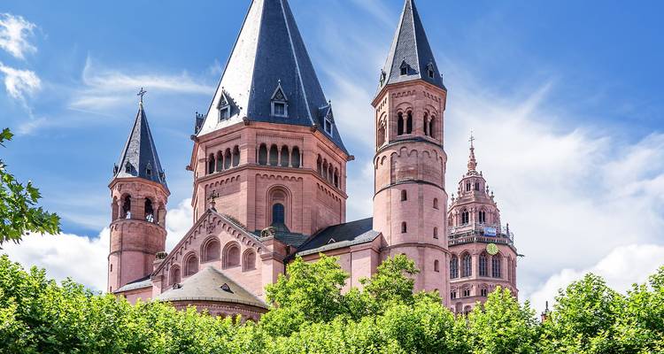 Primer plano de la Catedral de Maguncia de arenisca rosa con tres altas agujas enmarcadas por árboles verdes contra un cielo azul brillante.