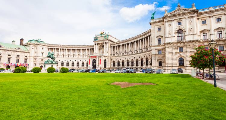 Vaste pelouse verte devant la façade néo-baroque imposante du palais de la Hofburg de Vienne sous des nuages épars.