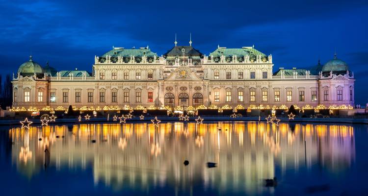 Iluminación nocturna del Palacio Belvedere con estrellas festivas reflejándose en su estanque ornamental.