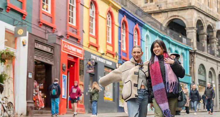 Dos mujeres caminando por una calle empedrada vibrante bordeada de escaparates multicolores en Edimburgo