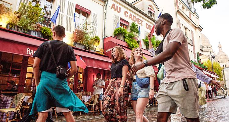 Groupe d'amis rit en marchant dans une rue pavée de Montmartre avec le Sacré-Cœur en arrière-plan.