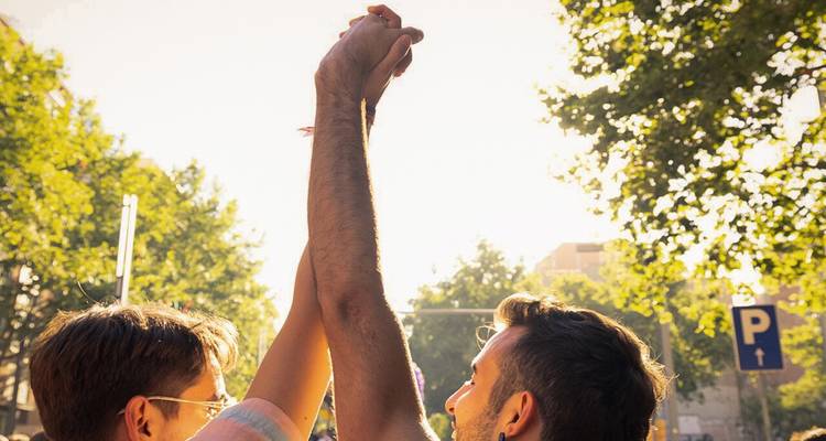 Dos hombres levantan las manos entrelazadas en celebración entre árboles urbanos iluminados por el sol en un evento al aire libre.
