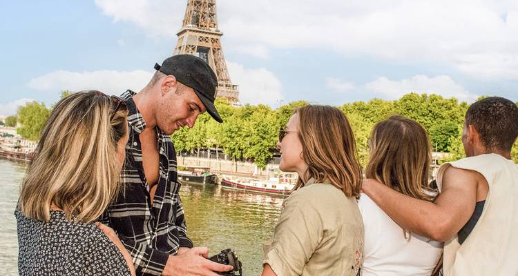 Grupo de viajeros conversando junto al río Sena con la Torre Eiffel alzándose detrás de ellos.