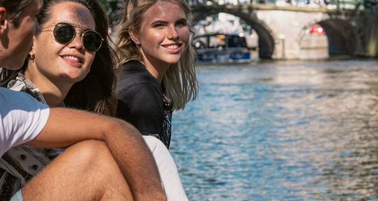 Close repeat image of friends seated by a city river with arched bridge and passing boats.