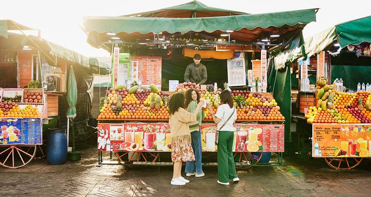 Amigos conversan frente a un colorido puesto de jugos marroquí lleno de frutas apiladas.