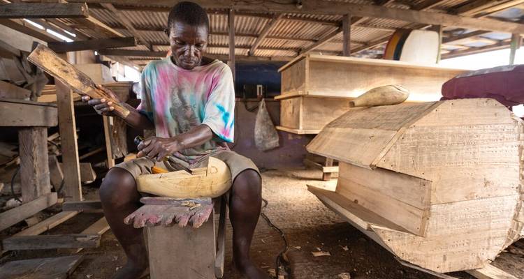 Een Ghanese ambachtsman snijdt een uitgebreid gevormde houten kist in een rustieke openlucht werkplaats gevuld met hout.