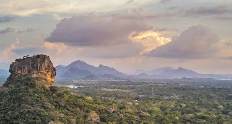 Dramatisch uitzicht op de Sigiriya Rots die boven dichte jungle uittorent met verre blauwe bergkammen onder pastelkleurige zonsondergangwolken.