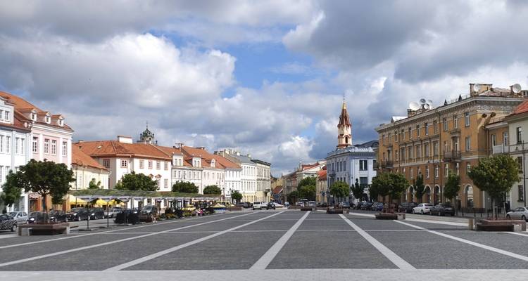 Plaza amplia y abierta rodeada de edificios históricos en tonos pastel y una aguja de iglesia bajo un cielo parcialmente nublado en Vilna.