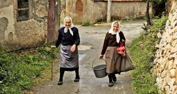 Two elderly women walking in a village road with a bucket and a sack.