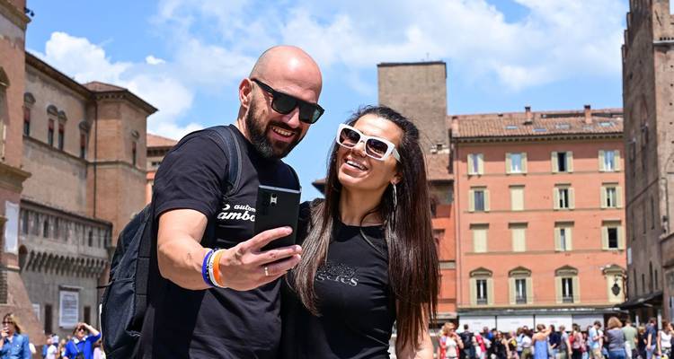 Jeune couple portant des lunettes de soleil prenant un selfie sur la place historique de Bologne avec des bâtiments en brique à tours en arrière-plan.