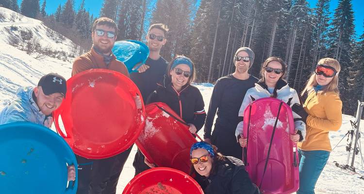 Un groupe d'amis tient des disques de luge lumineux sur une pente enneigée ensoleillée avec des sapins derrière.