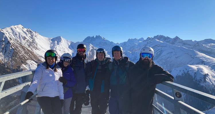 Six skieurs se tiennent sur une plateforme d'observation panoramique avec des pics alpins enneigés derrière eux.