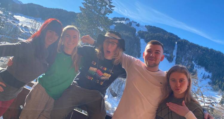 Cinq amis posent sur un balcon ensoleillé surplombant un village alpin enneigé et ses pentes.
