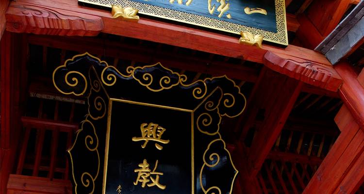Primer plano de una placa de madera ornamentada china con caligrafía dorada bajo los aleros del templo.