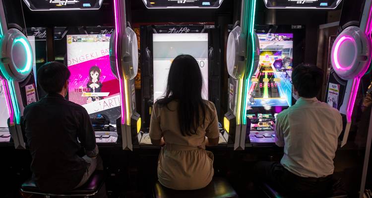 Tres personas sentadas frente a máquinas de juegos de ritmo brillantemente iluminadas en un centro de juegos japonés.