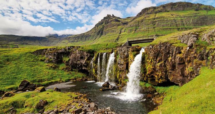Wasserfall, der eine felsige Klippe hinabstürzt, umgeben von grünen Hügeln.