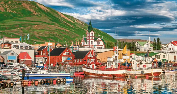 Buntes Hafendorf mit Booten und einer Kirche vor grünen Hügeln.