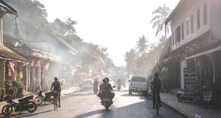 Rue poussiéreuse de fin d'après-midi à Luang Prabang remplie de cyclistes, de tuk-tuks et d'étals de marché baignés dans une lumière brumeuse.