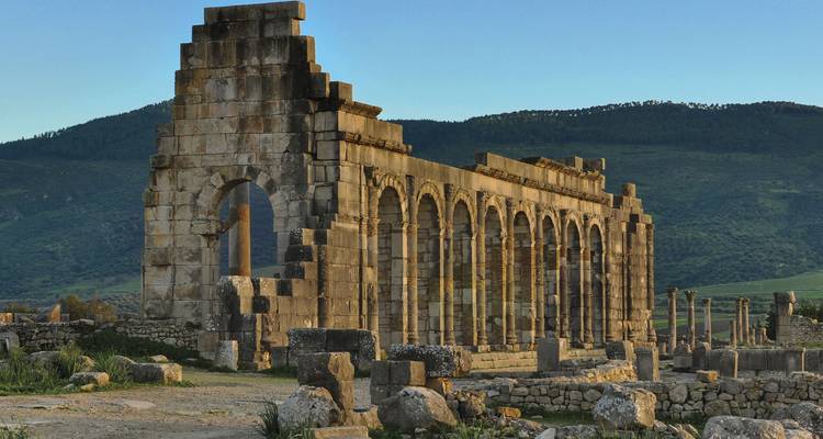 Arcada de piedra de Volubilis brillando al atardecer contra laderas boscosas.