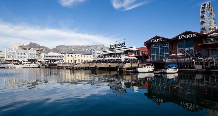 Escena pintoresca del V&A Waterfront con cielo azul, botes en el puerto y la Table Mountain de fondo reflejada en el agua.