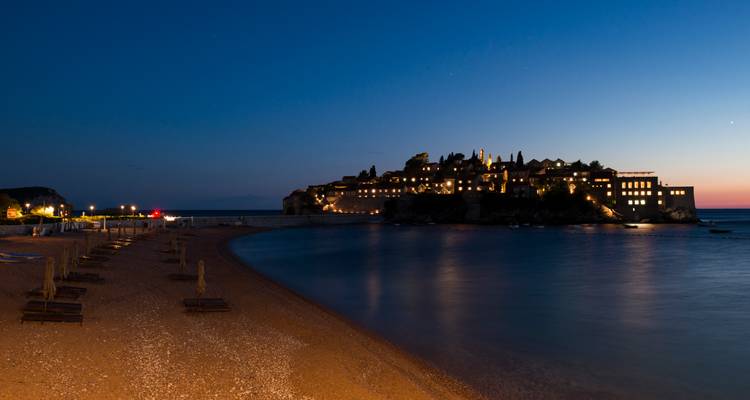 La isla resort Sveti Stefan brilla con luces al atardecer junto a las aguas tranquilas de la bahía.