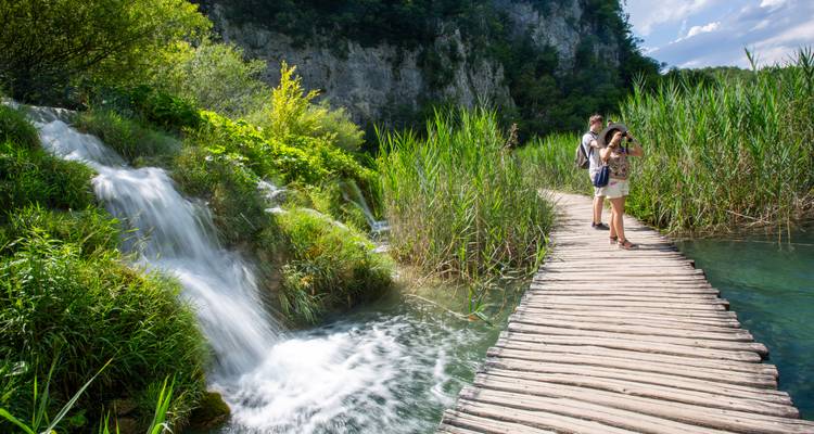 Twee jonge reizigers wandelen langs een rustieke houten promenade naast een kleine waterval met trappetjes omgeven door weelderig groen en hoge rietkragen.