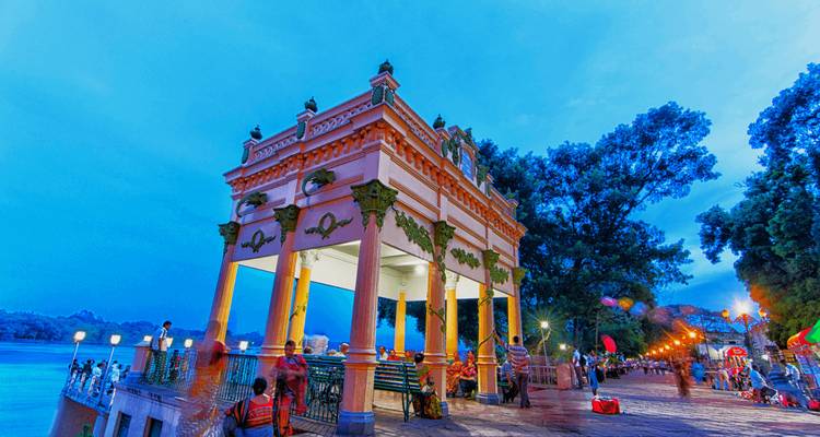Street scene at dusk with a pavilion and people walking along a riverbank.