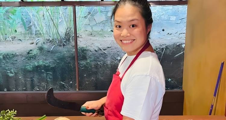 A smiling woman in an apron holding a knife indoors, with a natural backdrop.