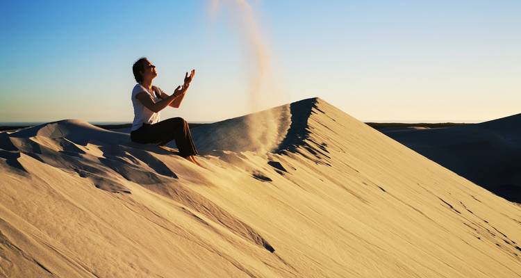 A person sits on a golden sand dune tossing a plume of sand into the air in late afternoon light.
