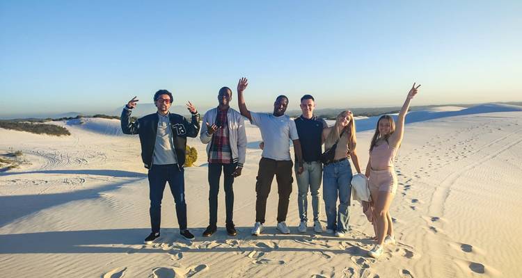 Six friends stand on sun-lit white sand dunes posing with peace signs and raised arms.