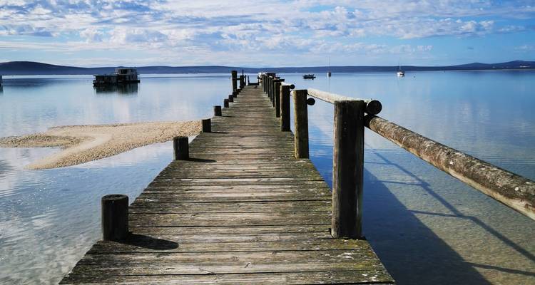 A weathered wooden jetty stretches over glassy blue lagoon waters under a lightly clouded sky.