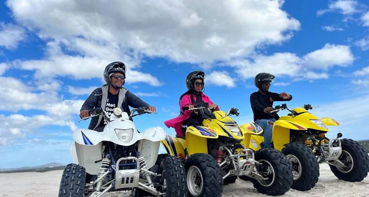 Three riders on brightly colored quad bikes pause on white sand beneath a vivid blue sky with puffy clouds.
