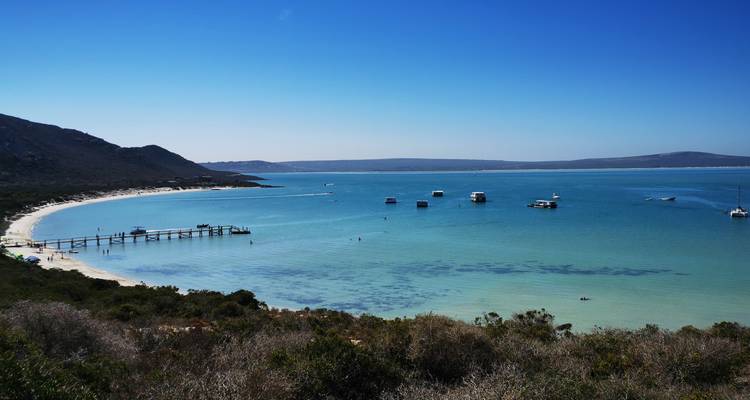 Panoramic view of a turquoise bay with a long pier and floating houseboats set against arid hills.