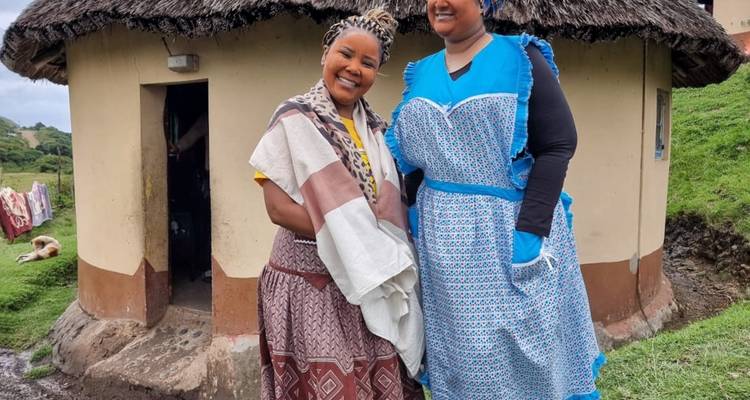 Deux femmes en robes traditionnelles à motifs sourient devant une hutte ronde au toit de chaume dans un village rural.