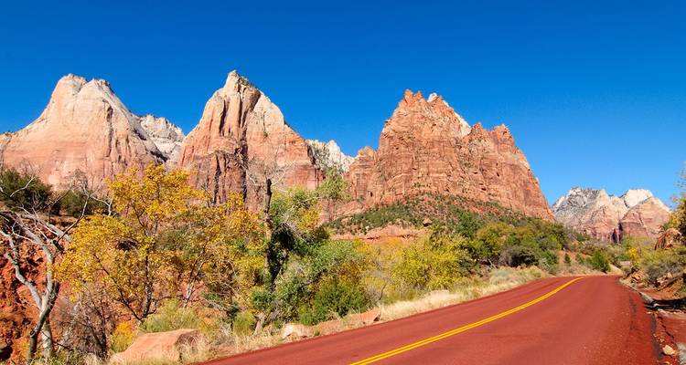 Una carretera de asfalto rojo se retuerce hacia acantilados de arenisca imponentes bajo un cielo azul impecable en el Parque Nacional Zion.
