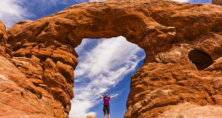 El excursionista se encuentra triunfante bajo un enorme arco de arenisca roja contra un vibrante cielo azul en el Parque Nacional Arches.