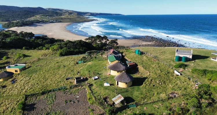 Aerial view of a rugged coastline with yellow rondavels on green hills above a sweeping beach and blue ocean.