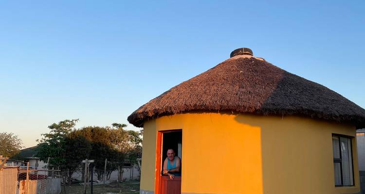 Traveller leaning in the doorway of a bright yellow rondavel under clear evening sky.