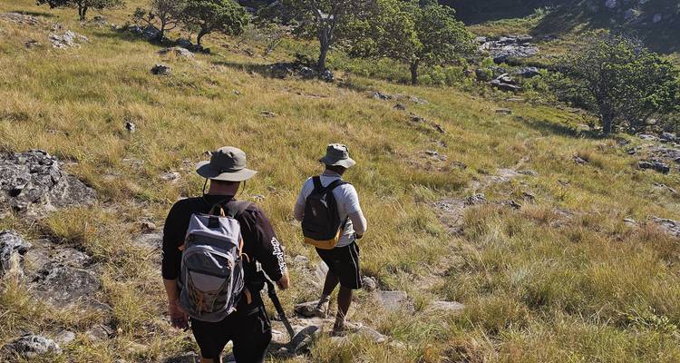 Two hikers with backpacks and hats walking along a grassy hillside trail.