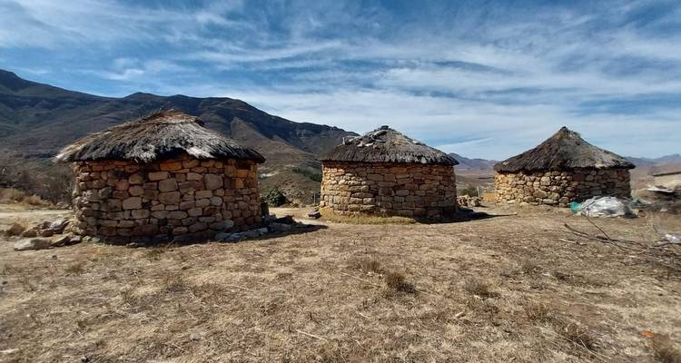 Drie traditionele stenen rondavels in een droog bergachtig landschap onder gestreepte wolken.